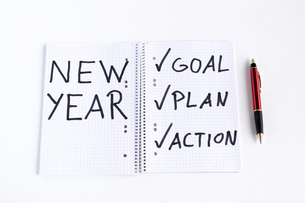 Notebook with writing of "New Year" and check marks beside Goal, Plan, and Action. With a red pen sitting beside the notebook.